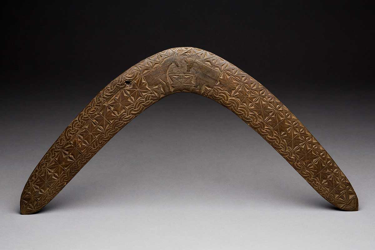 Studio photograph of a boomerang with incised intricate pattern and a carving of horse and cow at its apex.