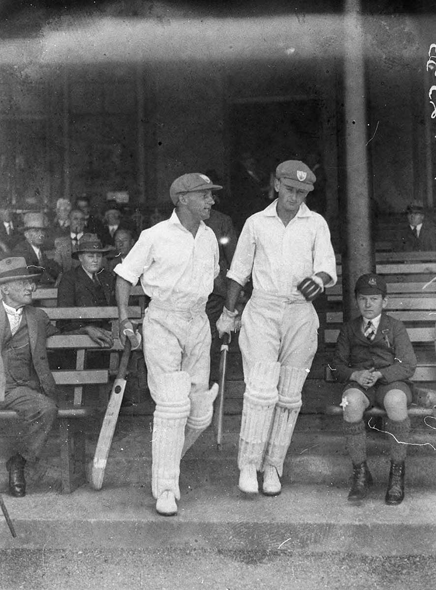 Two white-clad batsmen emerge from the stands with spectators on wooden benches on either side. - click to view larger image