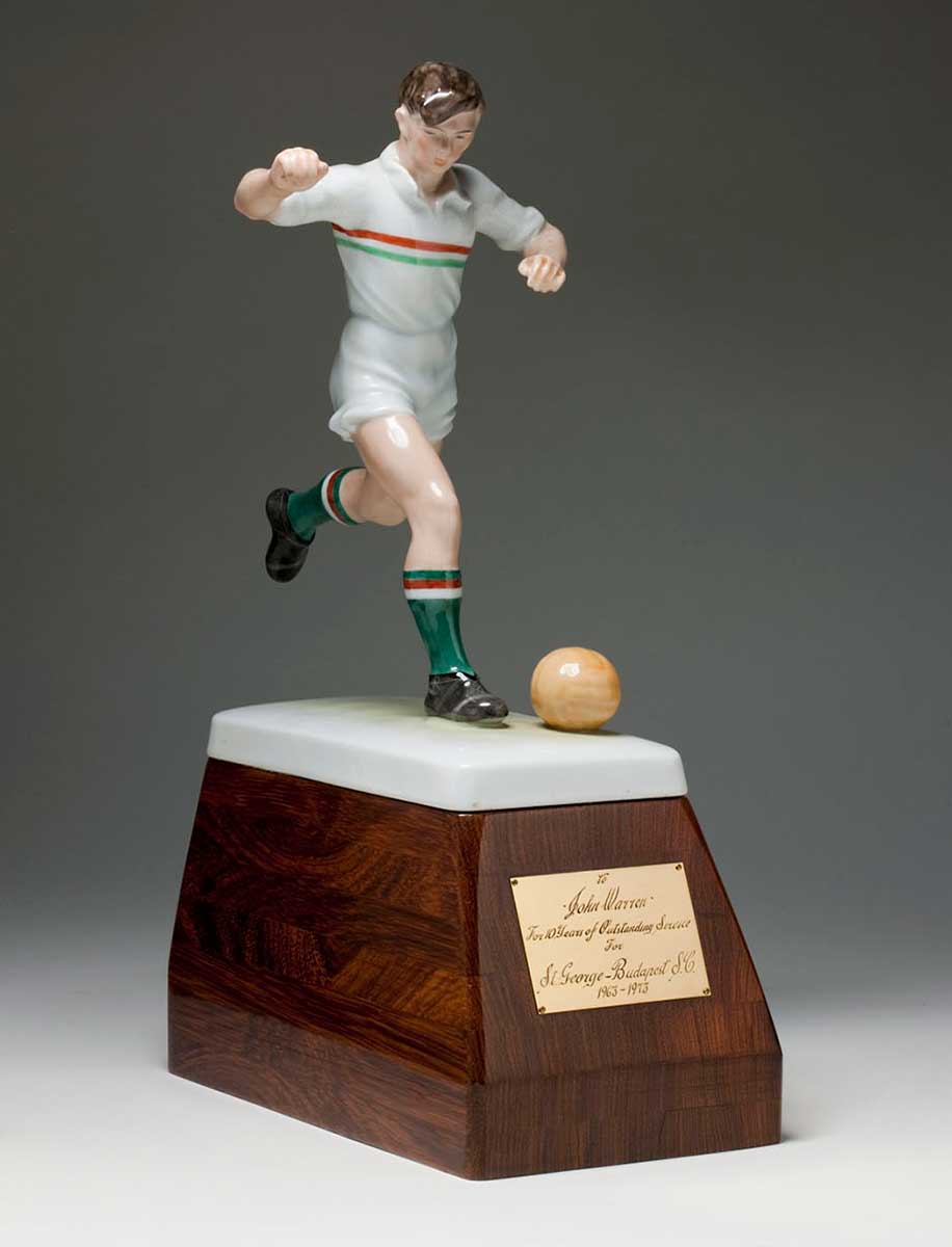 Ceramic figure of soccer player running up to kick a ball, with right leg on the ground and left leg extended backwards and bent at the knee. The painted figure wears a white uniform with a red and green vertical stripe across the chest, black boots and green socks with a red and white stripe. The figure is mounted on a wooden plinth with rectangular base and an engraved metal plaque at the front. - click to view larger image