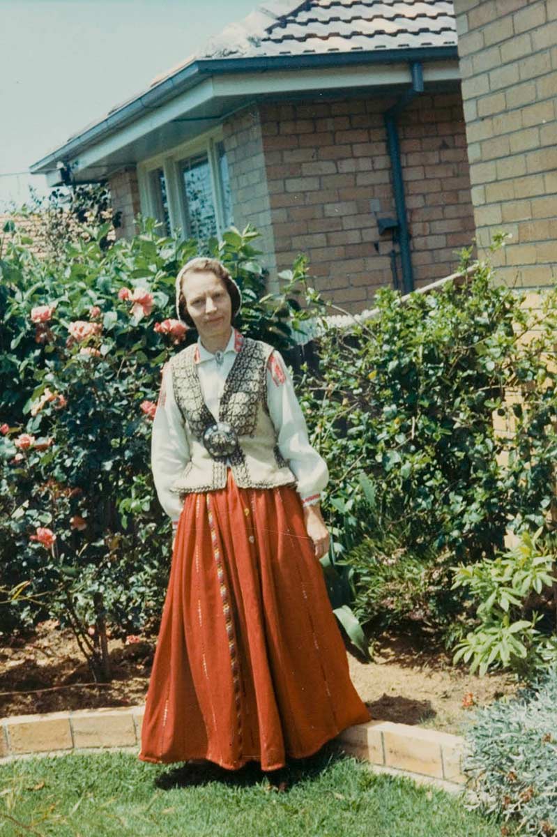 Colour photograph showing a woman dressed in Latvian national costume, consisting of a red skirt, beige waistcoat, white shirt and cloth cap. The woman stands in a suburban garden. - click to view larger image