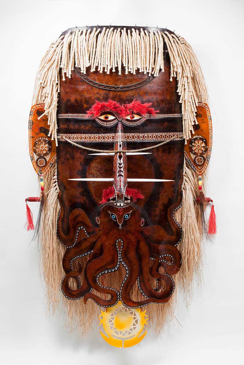 A brown coloured fibreglass mask with red plastic eyes. An octopus design is incorporated into the nose and mouth area with legs forming a beard. The ears are decorated with white linear designs and earrings made of beads and feathers. The nose is decorated with white linear designs, feathers and two nose ornaments. The hair is made from grass and rope. - click to view larger image