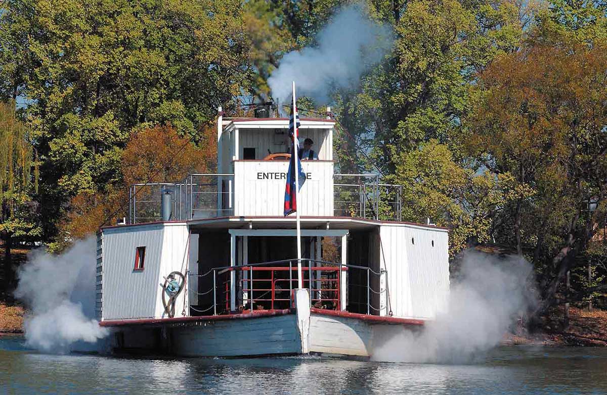 Paddle steamer on Lake Burley Griffin. A man stands in the 'ENTERPRISE' wheelhouse on the upper level. Steam can be seen coming from both sides of the lower deck of the white wooden vessel.