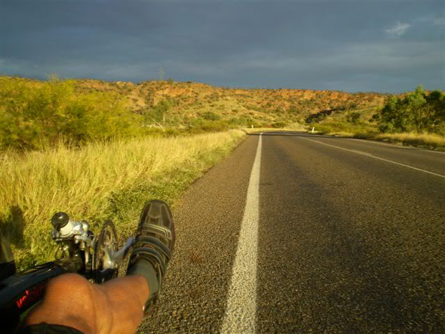 Colour photograph showing the front section of a recumbent bicycle and part of a man's leg, with this cycling shoe resting on a pedal. The bike is at the far left of a bitumen road, with a scrubby hill and cloudy sky ahead. - click to view larger image