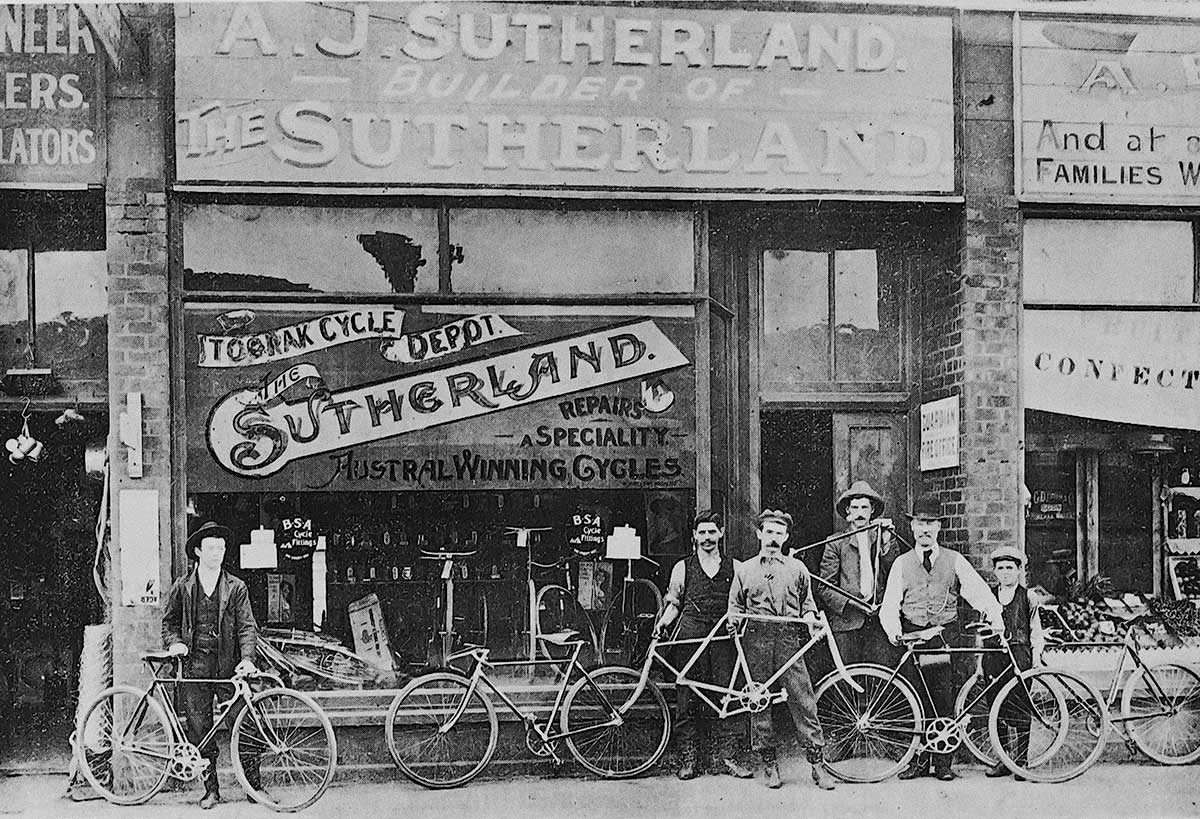Black and white photo of five men and a boy standing outside a shopfront with several bicycles. - click to view larger image