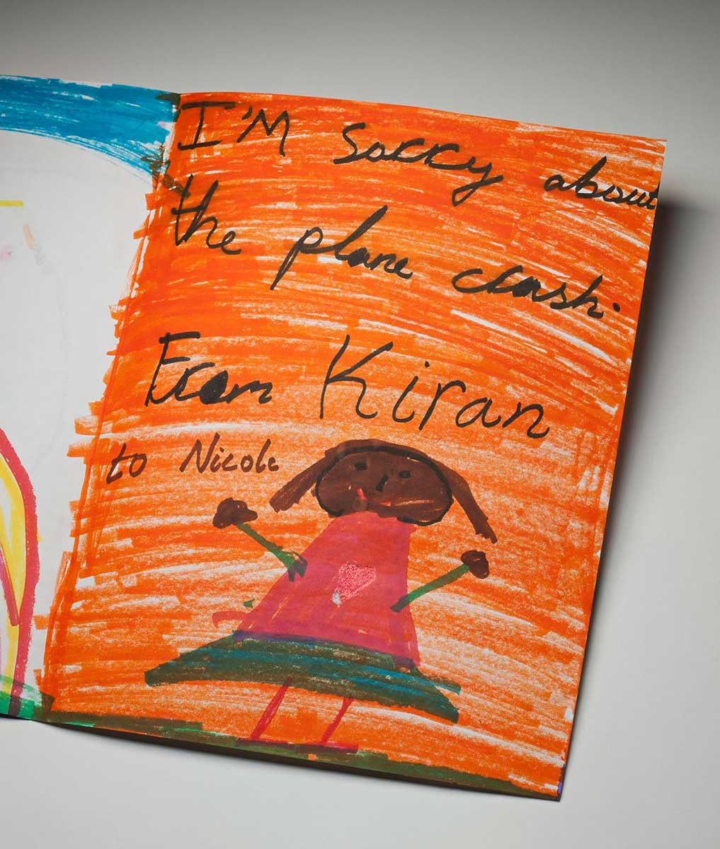 A handmade card showing the right inside view. The background is coloured in orange texta, with the handwritten words 'I'm sorry about the plane crash. From Kiran to Nicole' at top. There is a picture of a girl at the bottom of the card. She wears a pink top with a central heart motif, and a green skirt. She is smiling and her arms are stretched open. - click to view larger image