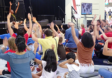 Woman sitting on a stage with audience of men, women and children sitting on the floor, raising their hands above their heads.