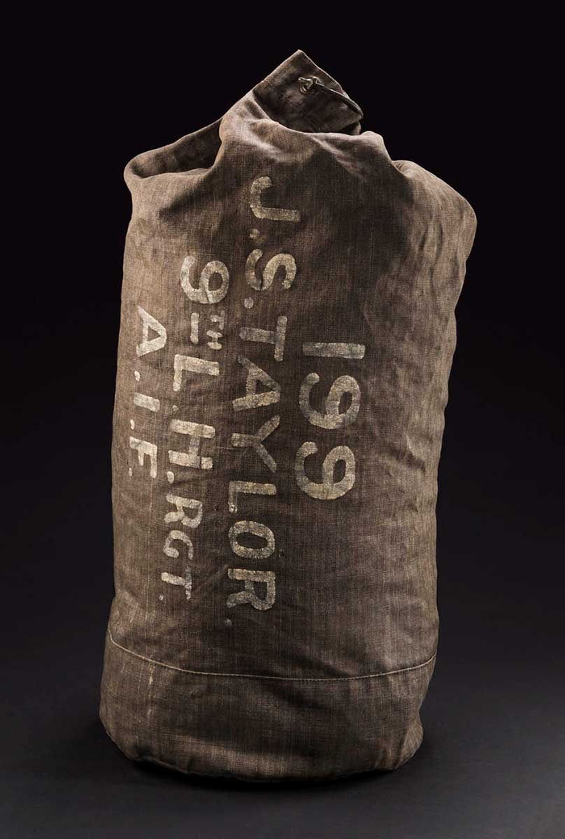 A dark coloured kit bag. It has a cylindrical shape with circluar holes at the top where a rope would have been threaded. One of the holes has a large metal clip through it. There is some writing in white paint on the side of the bag that reads '199 / J.S. TAYLOR / 9th L. H. RGT / A.I.F.' - click to view larger image
