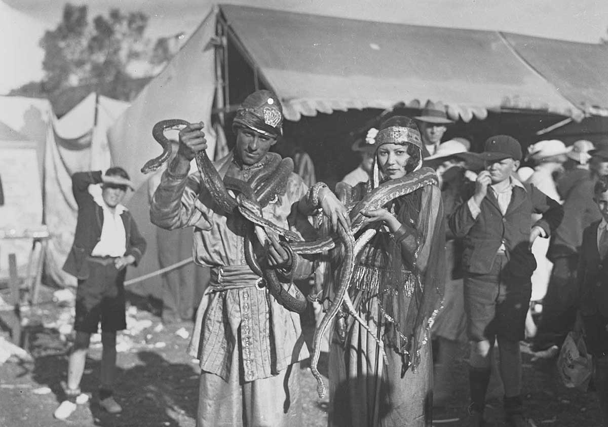 A man and a woman with snakes draped around their necks. Several onlookers are visible in the background. - click to view larger image