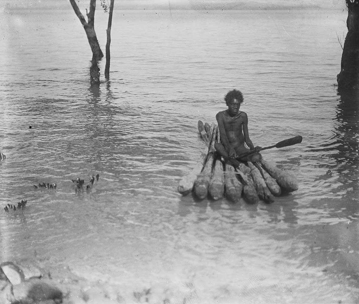 Black and white photograph of a man sitting on a raft on a body of water. - click to view larger image
