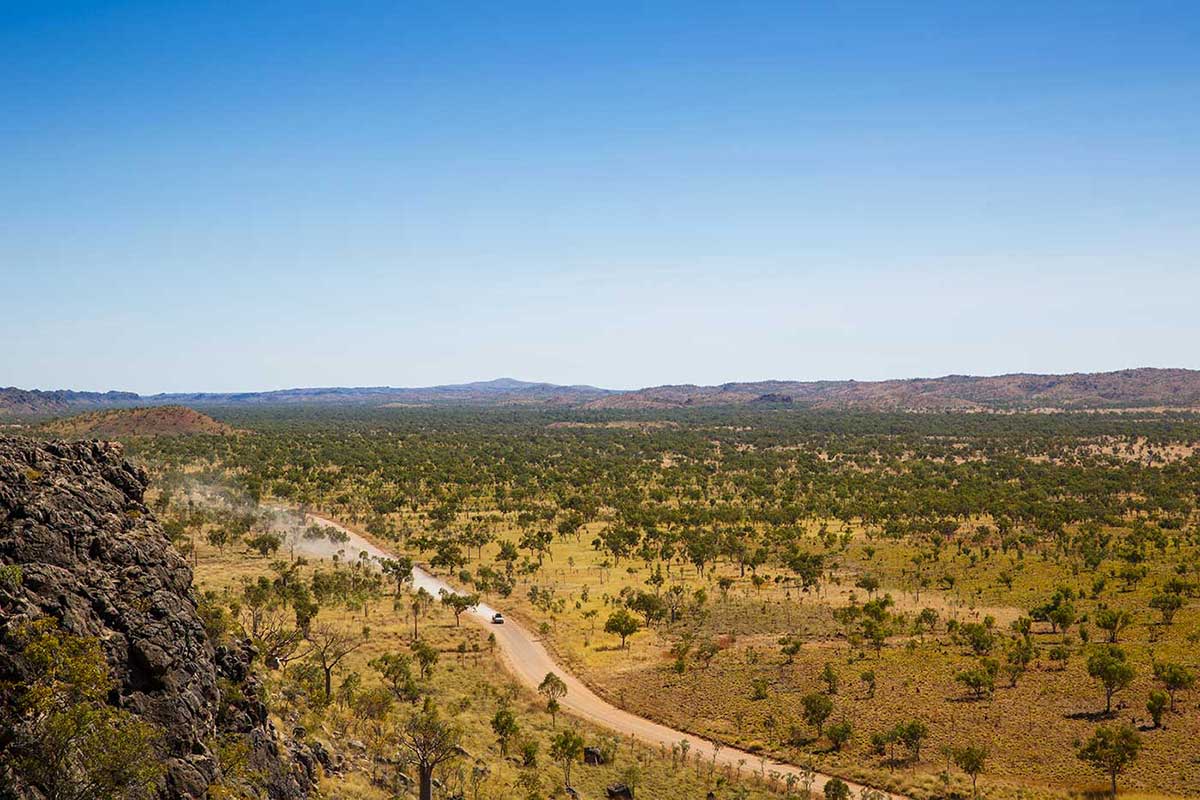 Colour photograph taken from a height and showing a dirt road winding through low, flat scrubland, with hills rising in the distance. - click to view larger image