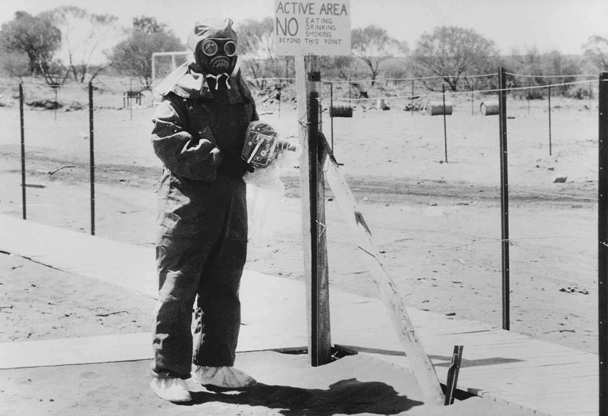 A man poses in dark cover-all with full headgear. A sign next to him reads ‘active area’.