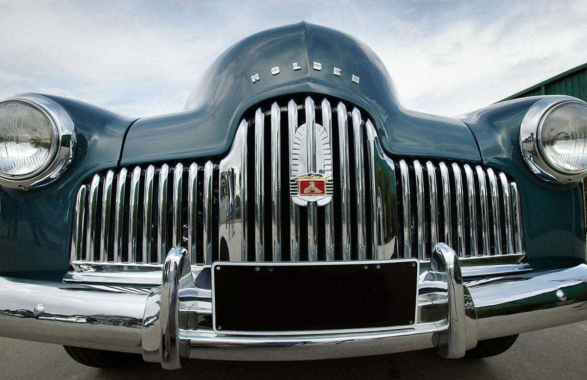 A blue-gunmetal grey sedan with chrome-plated bumper bar and radiator grille which bears a single Holden red lion badge with a small gold-coloured frame around it - click to view larger image