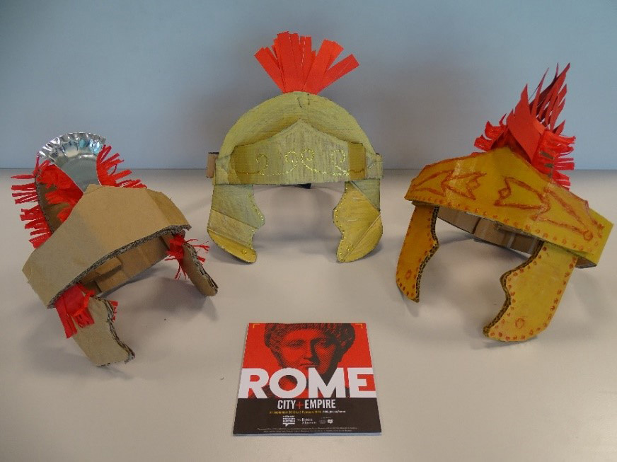 A photography of three handcrafted Roman helmets. A book with the title in large white letters, ROME, sits in the middle of the display.