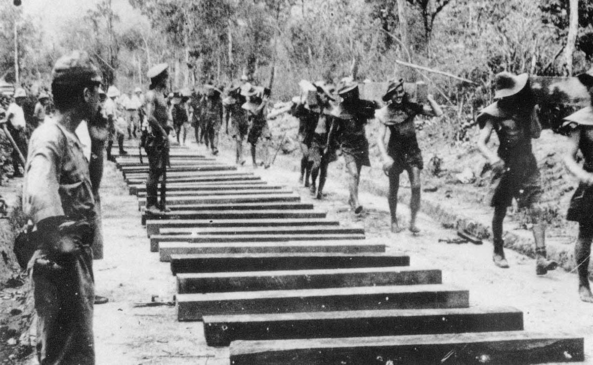 Black and white photograph of men carrying railway sleepers, and being watched by soldiers. - click to view larger image