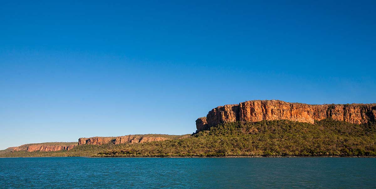 A photo of craggy cliffs overlooking a bright blue sea. The sky is a matching colour of bright blue and underneath the cliffs are dense bushland.