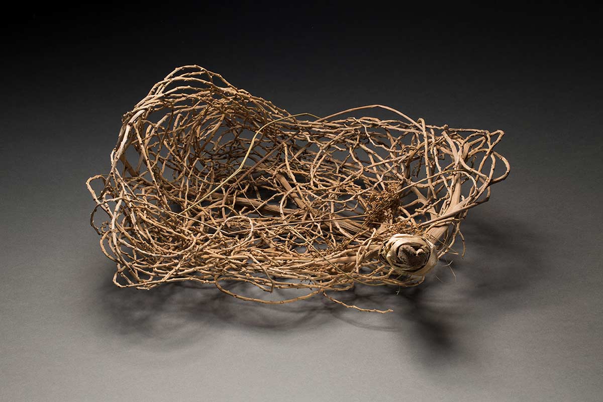 An oval shaped woven basket made from bangalow palm stalks. There are three stems of bangalow palm seeds woven loosely to the basket near the binding on the main branch. - click to view larger image
