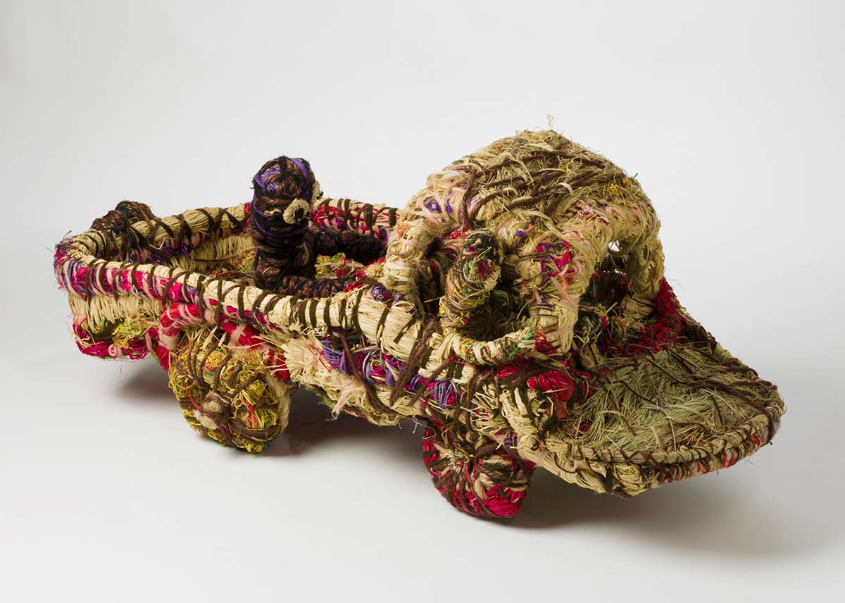 A sculpture depicting a truck with two people in the cabin and one person travelling on the back . The sculpture is made of grass with woven plant material dyed in different colours and strands of wool. - click to view larger image