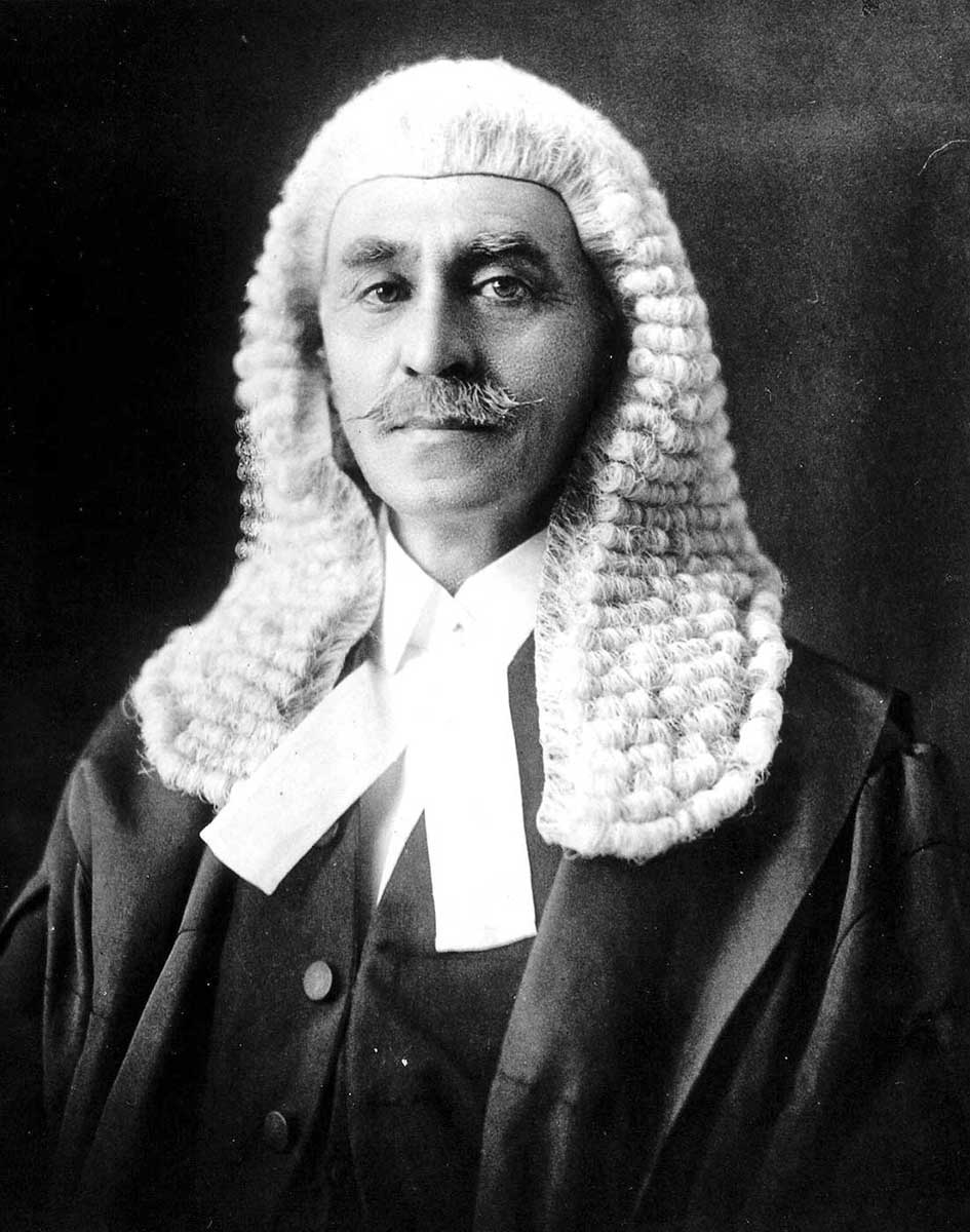 Studio portrait late middle-aged man, unsmiling with greying pointy moustache wearing judge’s wig and robes.