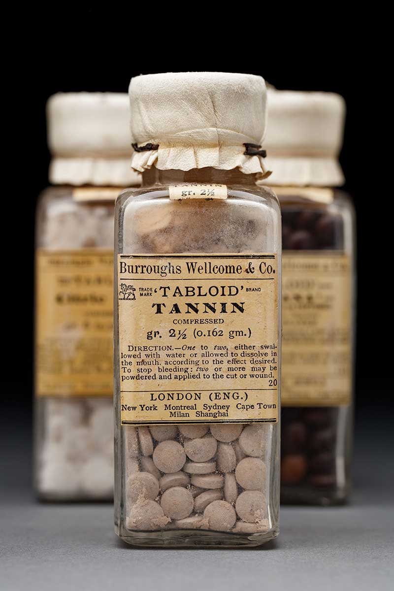 Three glass bottles filled with small, round pills. The bottle at the front has a printed label that starts, 'Burroughs Wellcome & Co, Trademark 'Tabloid' brand Tannin.'