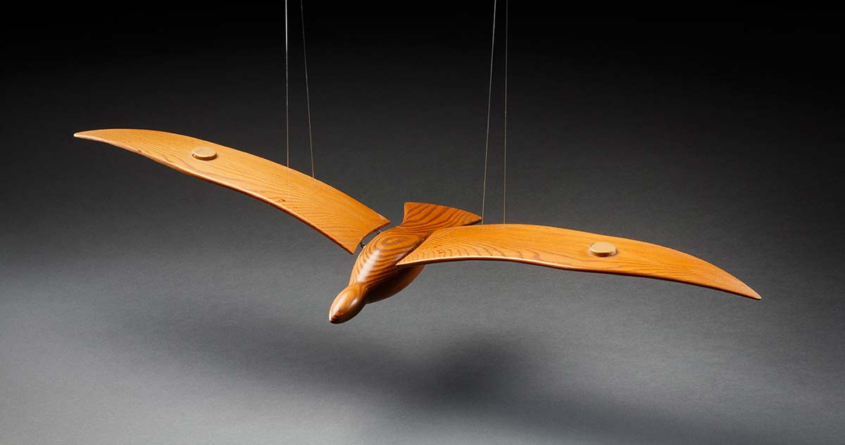 A carved wooden seagull with adjustable wings, hung as a mobile. The bird is made from three pieces of timber which have a strong grain pattern. The wings are attached with metal fixings, and the hanging device is made from a clear plastic tube and multiple sections of clear mono-filament. - click to view larger image