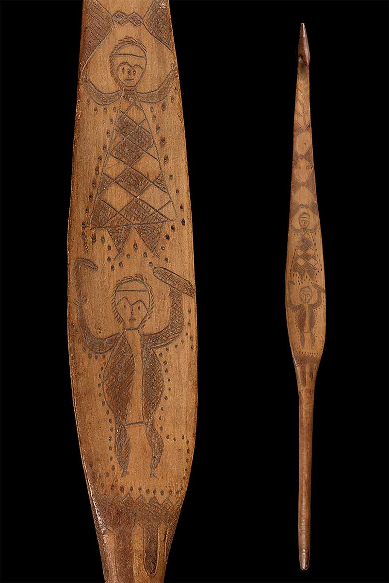 Spear-thrower made of wood, decorated with complex incised figurative designs, including two human figures; one holding a shield and a boomerang, the other clothed in a decorated skin cloak. - click to view larger image