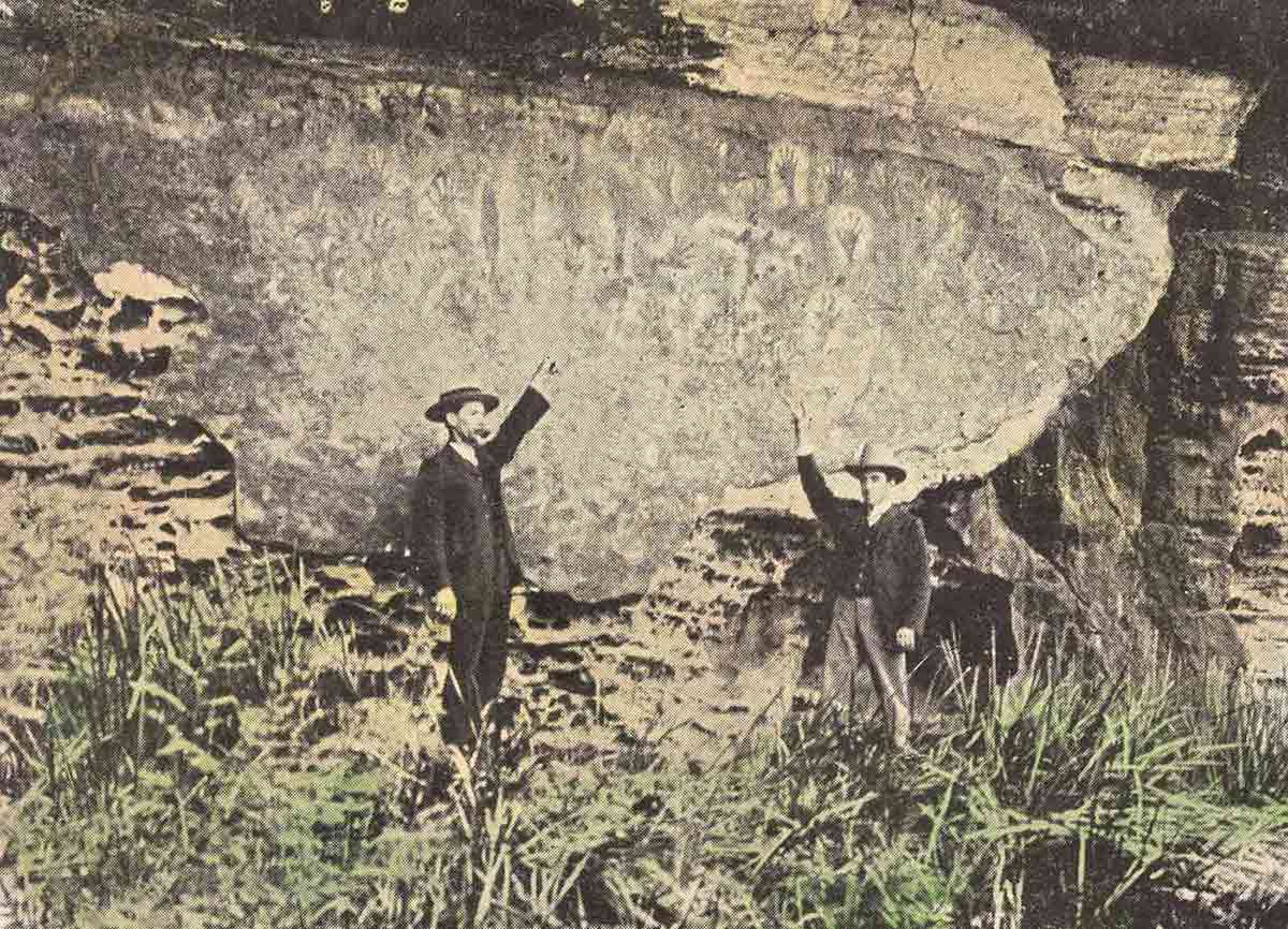 Postcard showing two men pointing up at hand stencils on a rock face. - click to view larger image
