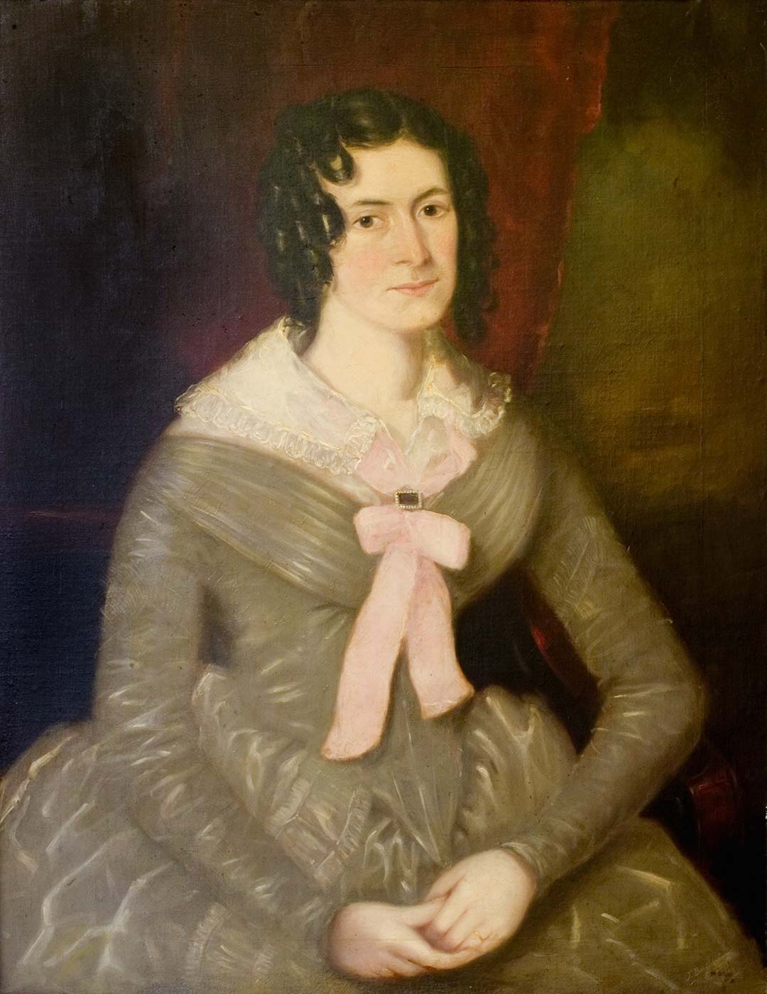 Portrait of a lady in 1845 seated with her hands folded together in her lap, with ringlets around her face wearing an olive-coloured dress with full skirt, tight waist and lace collar tied with pink bow and clasp at the front. - click to view larger image