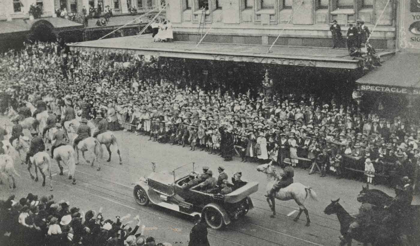 Black and white photo of a procession down a city street featuring horse and riders leading an automobile which is carrying several people, while crowds look on. - click to view larger image