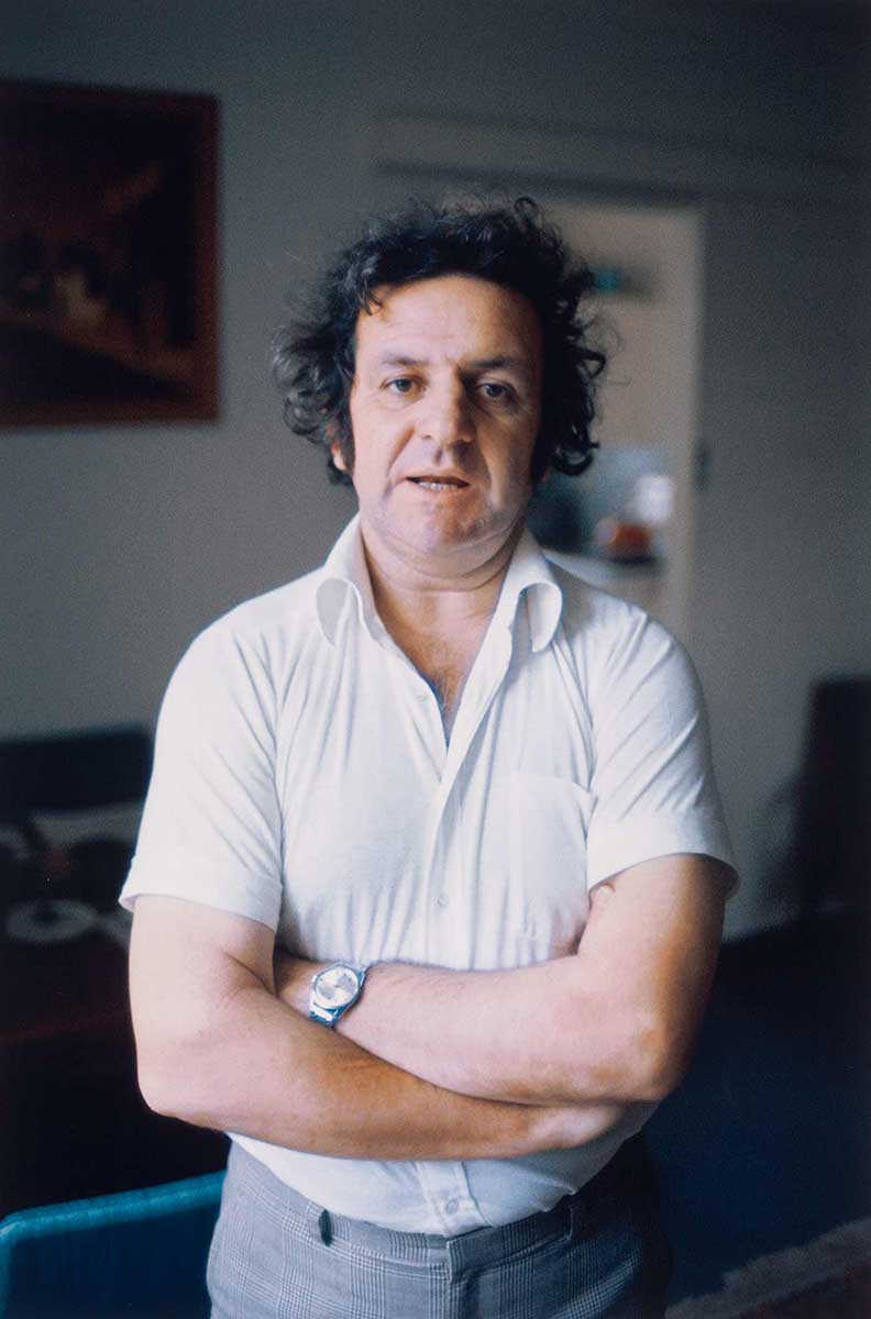 A man wearing a white polo shirt and grey trousers is looking at the camera and standing with his arms crossed. - click to view larger image