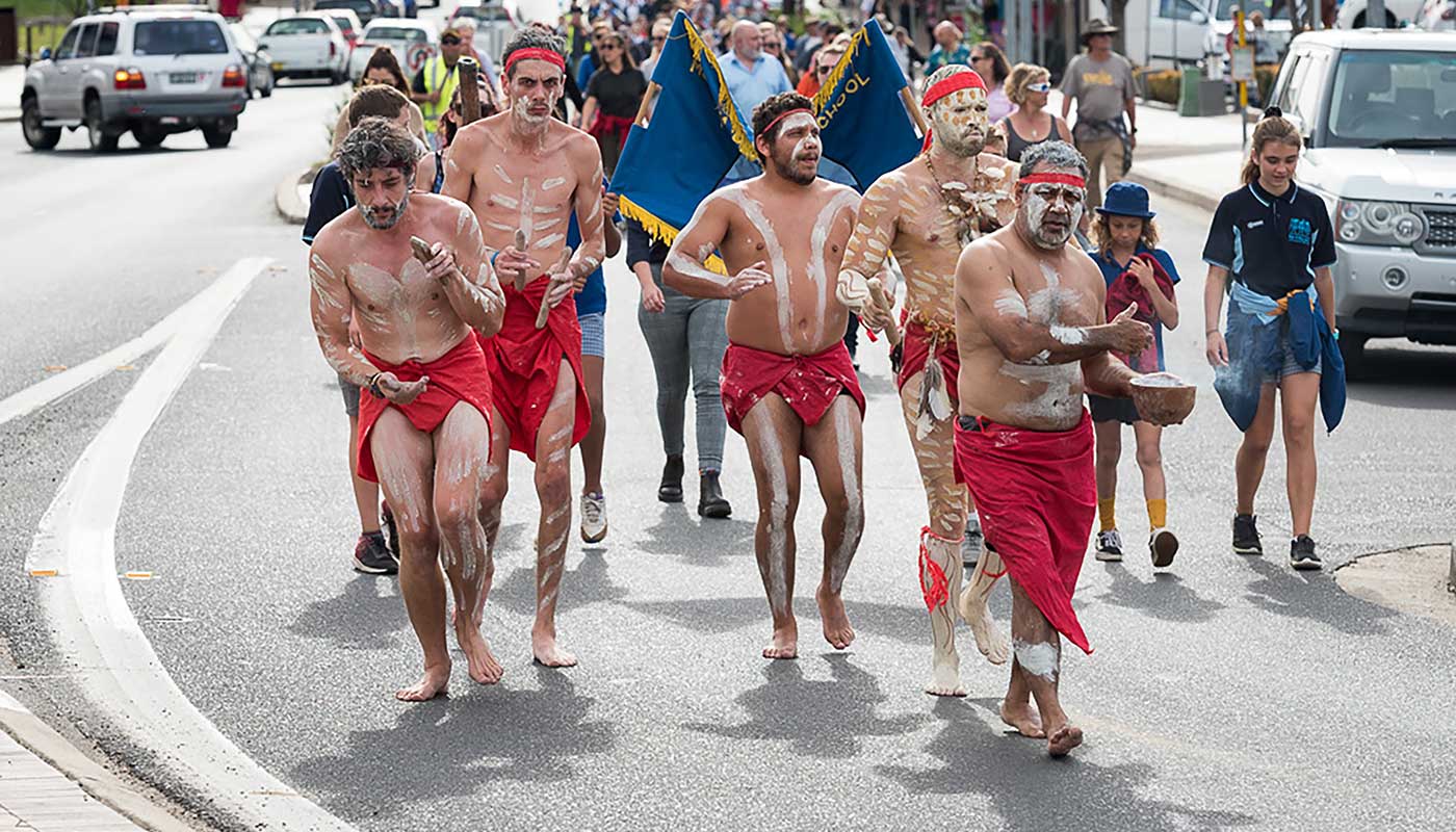 On an annual Sorry Day walk, a small group of people adorned with body paint lead a larger crowd on a busy road in Eurobodalla.