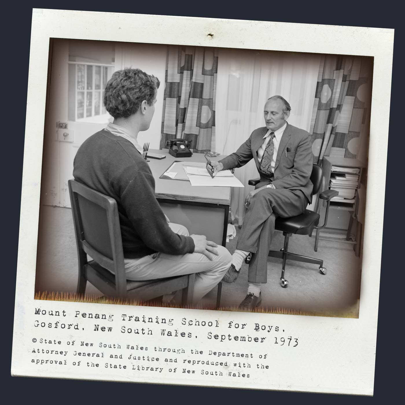 A black and white Polaroid photograph of an older man and someone who appears to be an adolescent boy sitting in an office. The man is dressed in a suit and tie and has his left leg crossed over his right. He is holding a pen and looking at a document on his desk. The boy is seated facing him with his right hand placed above his knee. He appears to be wearing a dark coloured jumper and light coloured trousers. Typewritten text underneath reads 'Mount Penang Training School for Boys, Gosford, New South Wales, September 1973.' Below this text is the circle c copyright symbol followed by 'State of New South Wales through the Department of Attorney General and Justice and reproduced with the approval of the State Library of New South Wales.' - click to view larger image