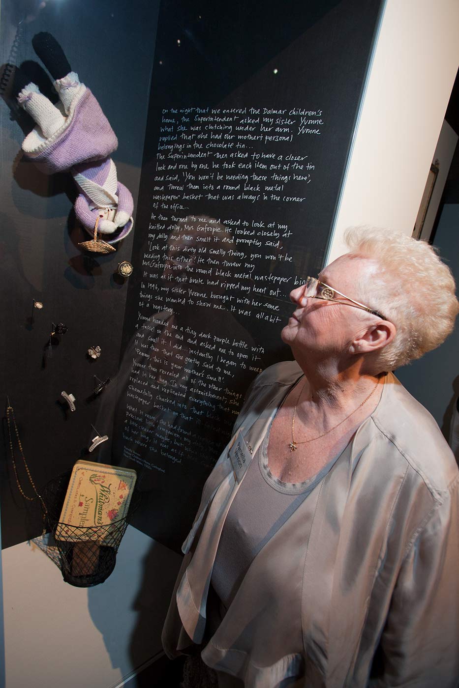 Colour photograph showing a woman standing, looking into an exhibition case. On the back wall of the case a small knitted doll tumbles down towards a metal wastepaper basket, which has a 'Whitman's Sampler' chocolate tin inside. Small hair clips and a pair of rosary beads are mounted between the doll and the bin. White handwritten text on a dark background is visible at the left of the case. - click to view larger image