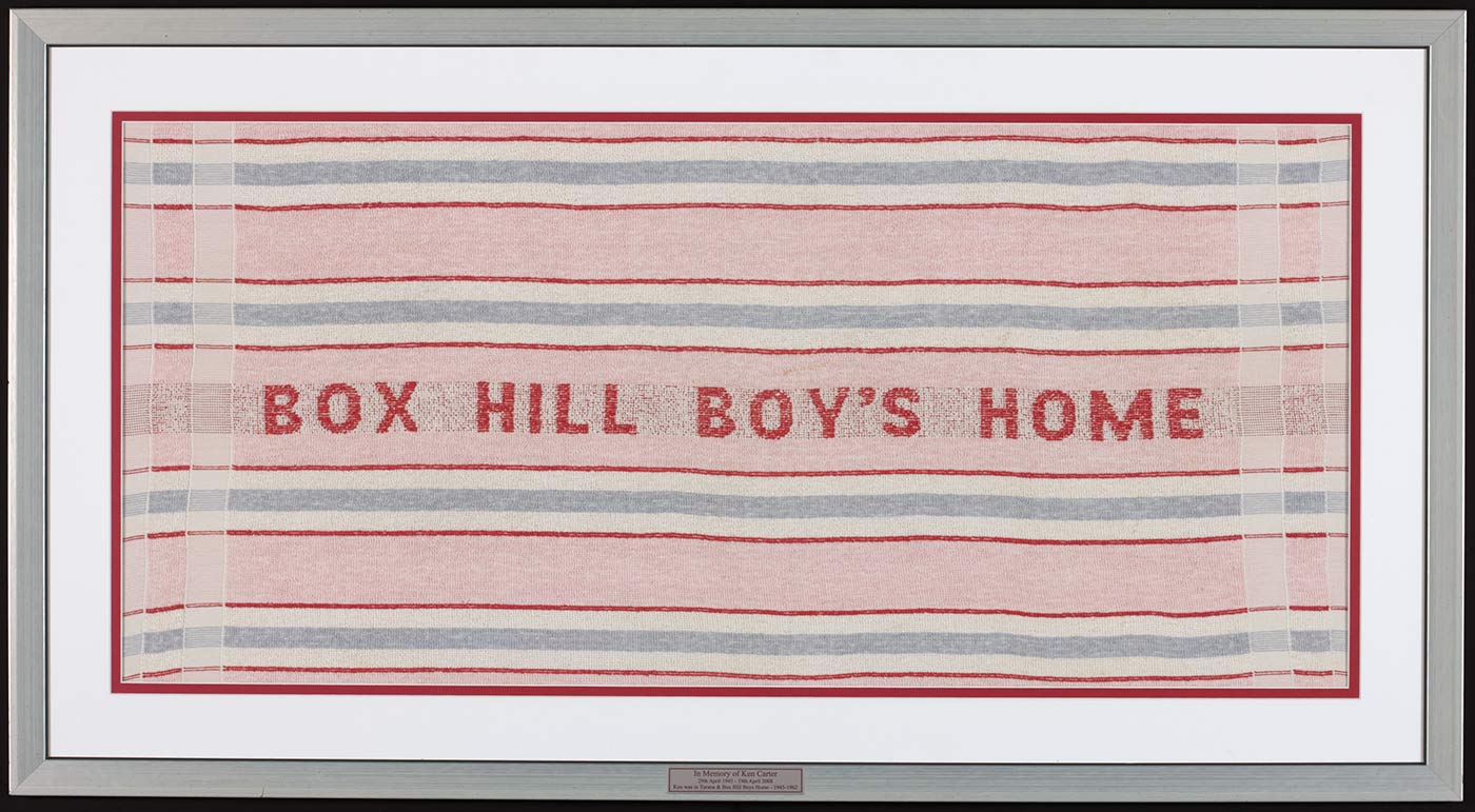 Photograph of a red, white and blue striped towel set in a silver coloured frame. The towel has 'BOX HILL BOY'S HOME' printed in red capitals at the centre. A small plaque, bottom centre of the frame, reads: 'In Memory of Ken Carter / 29th April 1945 - 19th April 2008 / Ken was in Turana & Box Hill Boys Home 1945-1962'. - click to view larger image
