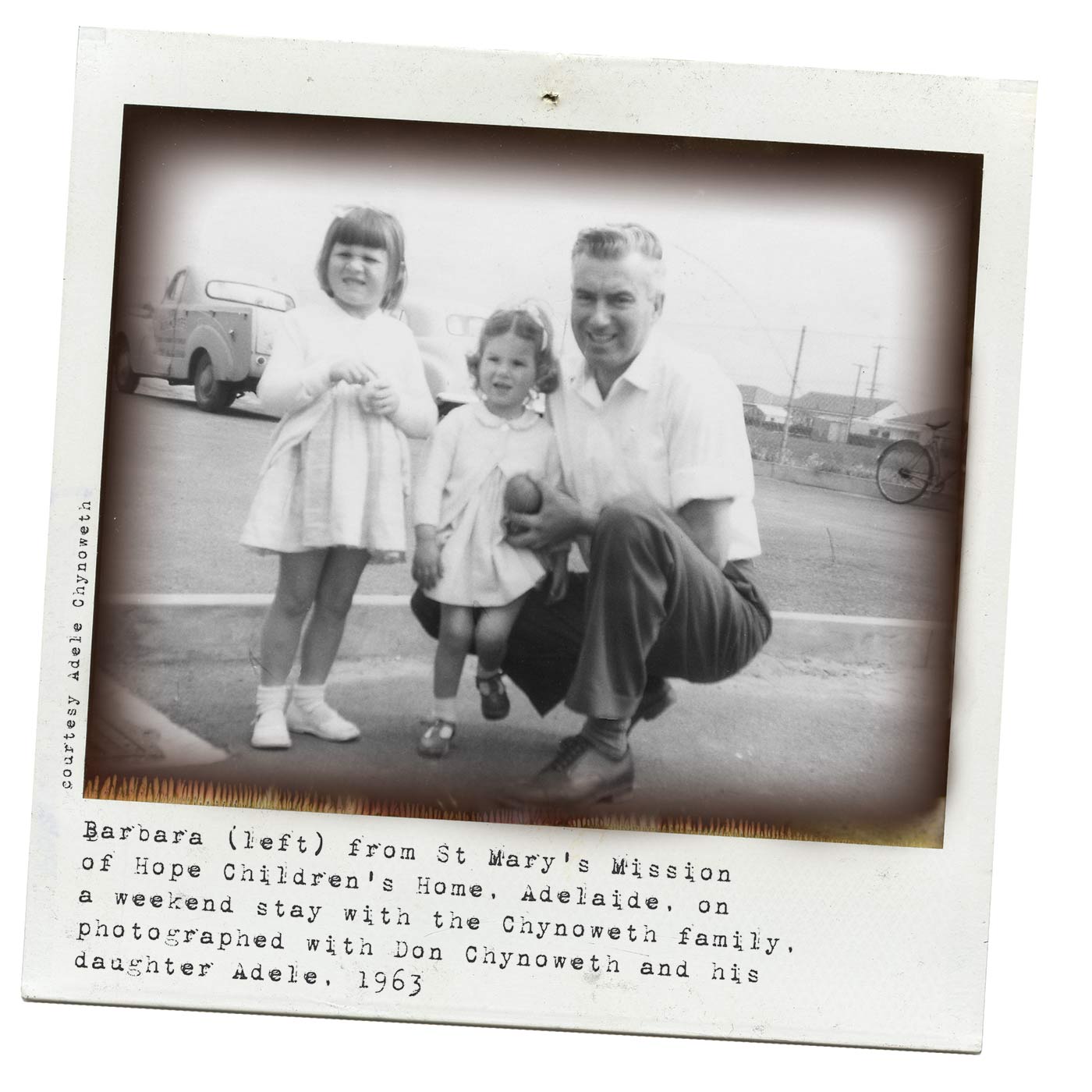 A black and white Polaroid photograph of a man with two little girls. The man is crouched down to be at the same level as the girls and all are smiling at the camera. Typewritten text underneath reads 'Barbara (left) from St Mary's Mission of Hope Children's Home, Adelaide, on a weekend stay with the Chynoweth family, photographed with Don Chynoweth and his daughter Adele. 1963'. Smaller text, on the left-hand side of the image, in a vertical direction, reads 'Courtesy Adele Chynoweth'. - click to view larger image