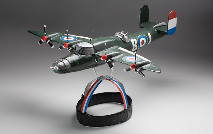 An aeroplane headdress modelled on a British Mitchell Carrier plane, circa WWII. The metal headpiece is painted in red, white and blue stripes, with black adhesive tape wrapped around the outer band. On top of the metal headpiece, is a painted wooden model of a British Carrier Plane, possibly made of balsa wood, supported by metal struts under the wings. The aeroplane is painted green with markings in blue, red, white and yellow. The wings each feature a target or bullseye design, and two propellers.