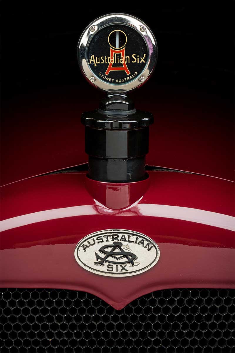 View of a polished red motor car bonnet and badge that reads ‘Australian Six’. - click to view larger image