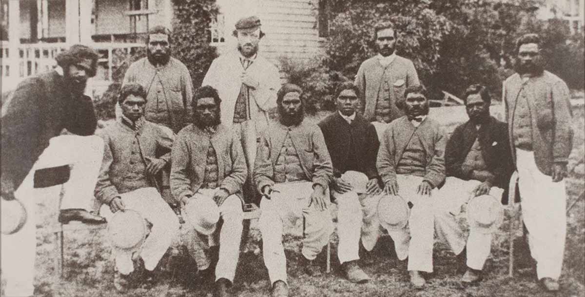 Black and white reproduction photograph of a cricket team of eleven men, six of whom are seated. They are outside in front of a building.