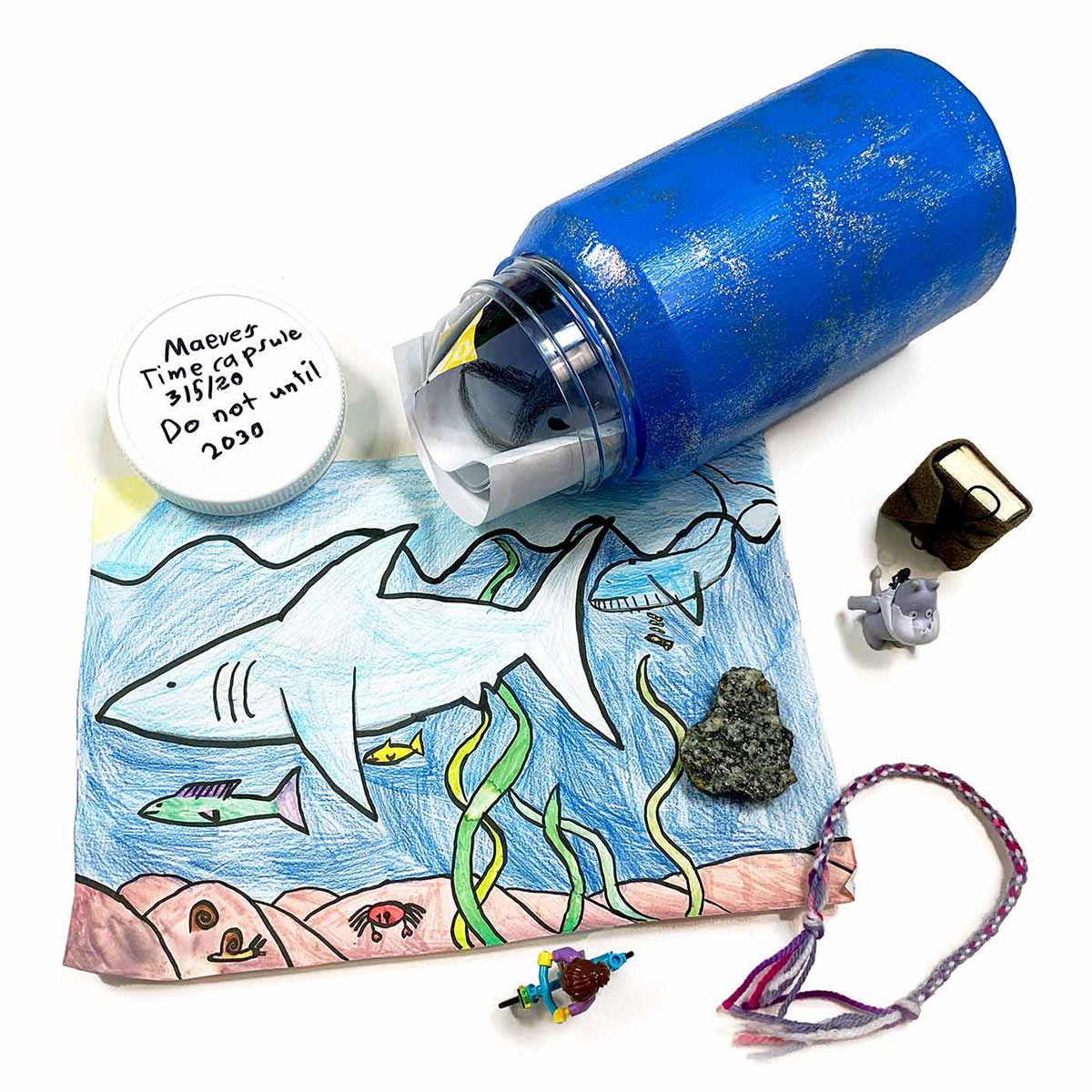 Time capsule featuring a bottle with folded drawings on paper inside. The bottle is sitting next to other objects including a miniature diary, a Lego person on a bike, and a rock. These objects are sitting on top of a drawing featuring an underwater ocean scene. On top of the bottle’s cap is written in black marker: “Maeves Time capsule. 3/5/20. Do not until 2030”. - click to view larger image