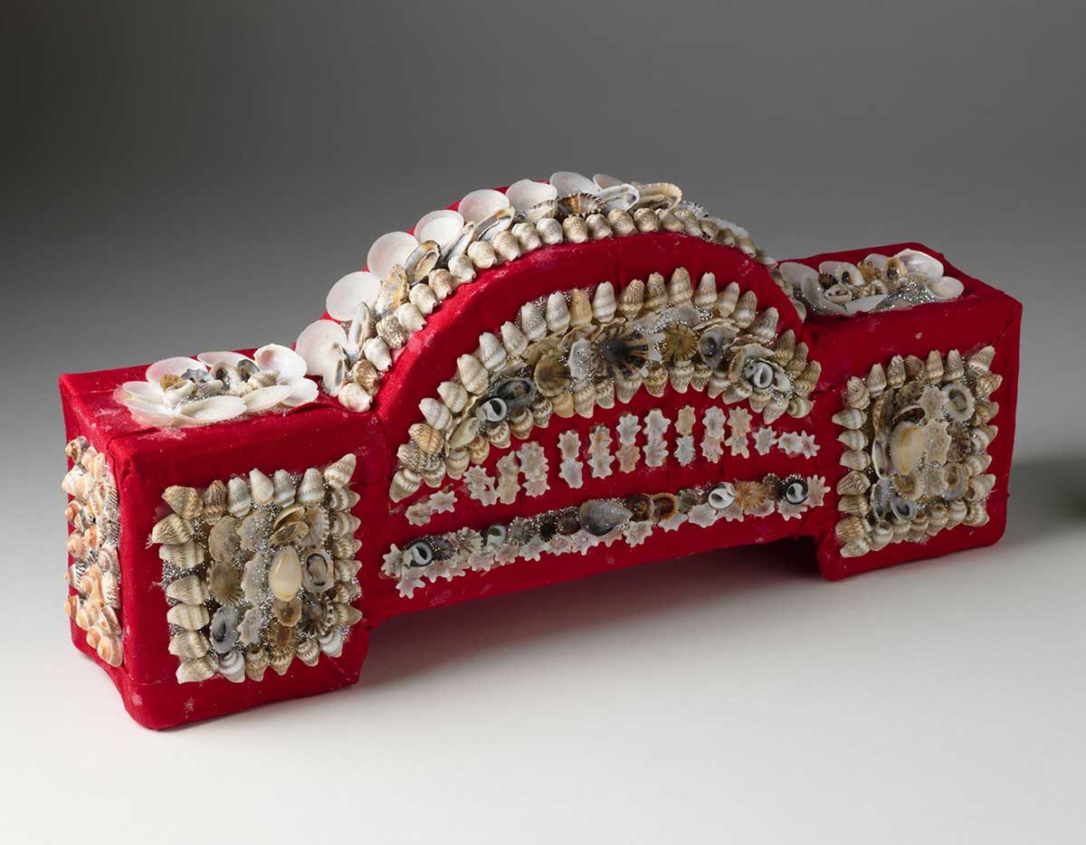 Ornamental shellwork Sydney Harbour Bridge. Several kinds of small beige shells are glued in rows to form blocks on a red cotton fabric-covered formwork, which is shaped like the Sydney Harbour Bridge. - click to view larger image