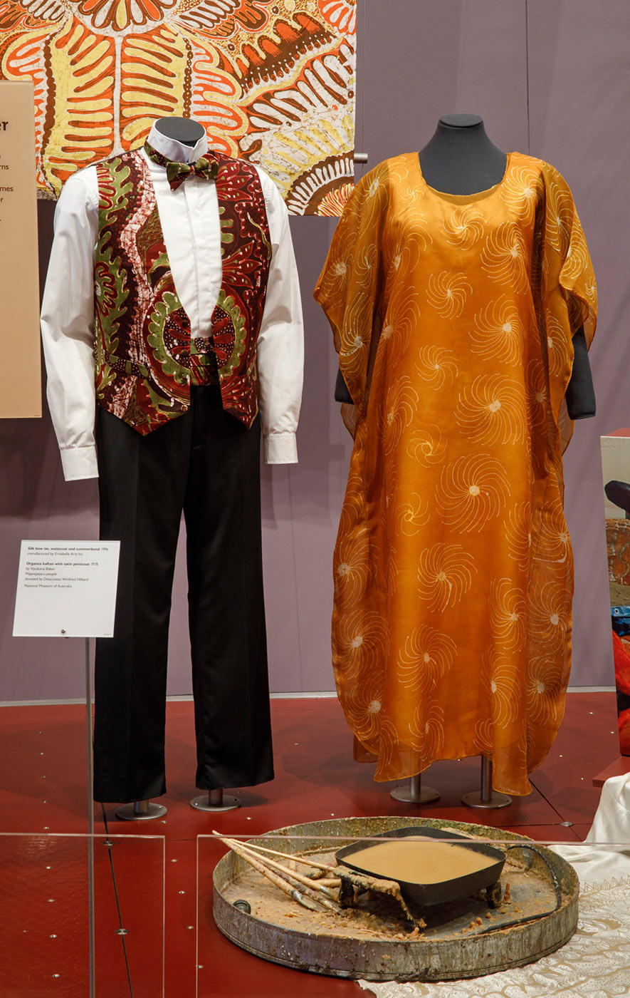 Museum display showing two mannequins. On the left, one has black pants, white shirt and a brightly coloured silk waistcoat. The second wears a bright orange kaftan. - click to view larger image