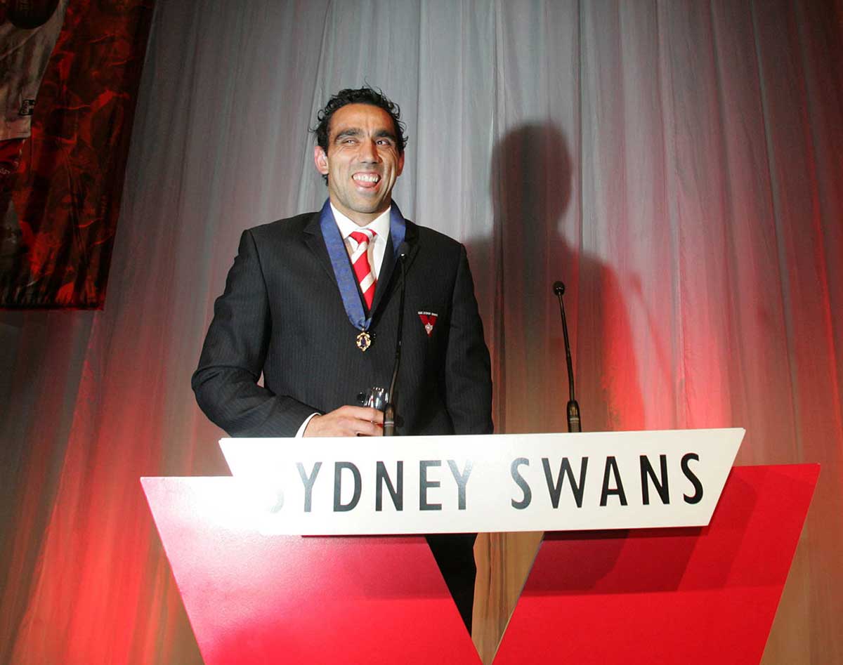 Colour photo of Adam Goodes on a stage behind a Sydney Swans sign. He is smiling broadly and wearing the Brownlow medal. - click to view larger image