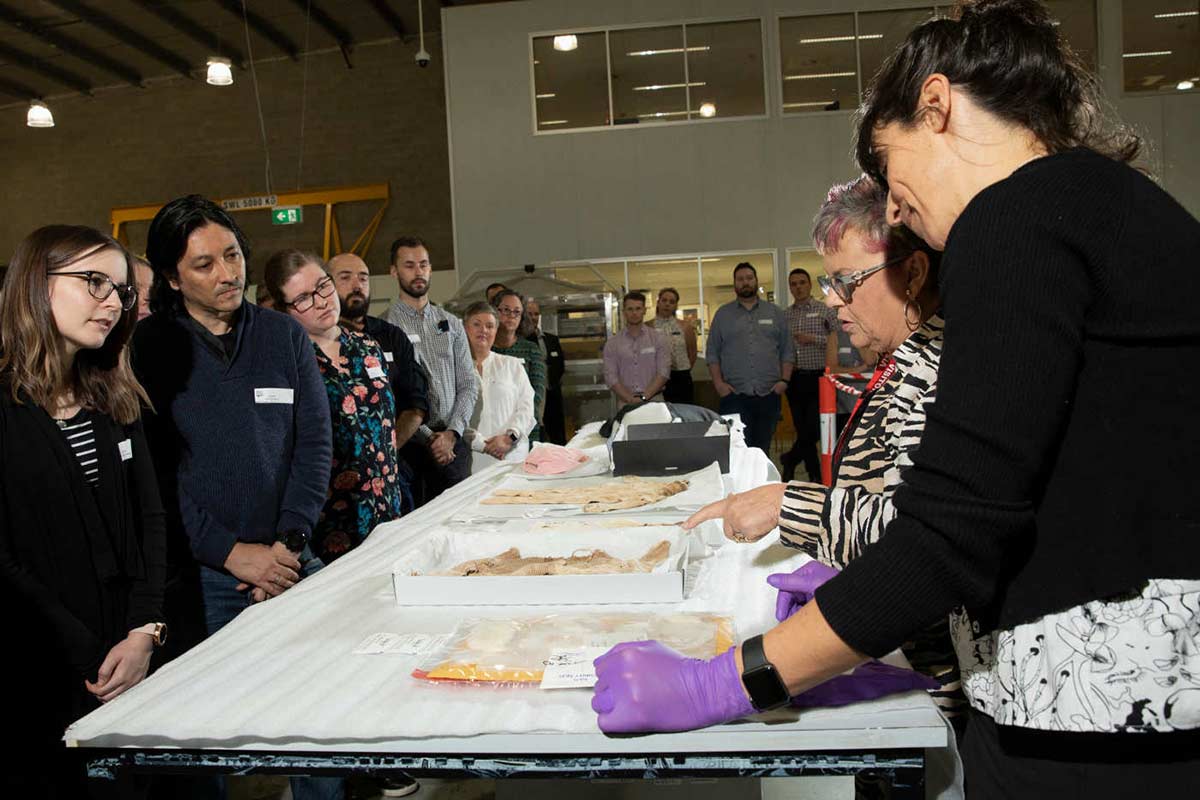 Two women stand behind a high table, looking down at several pieces of clothing on display. A group of visitors stand on the other side and at the end of the table, watching. - click to view larger image