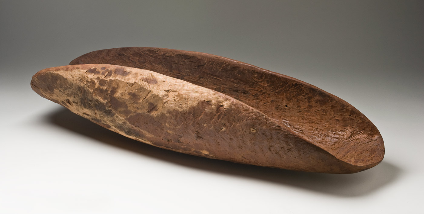 A red-brown wooden concave oval container with dark patches on the sides of the outer surface. The inner surface is rough with the tool marks and there are short parallel grooves along one end of the inner surface. The outer sides are a light beige colour. - click to view larger image