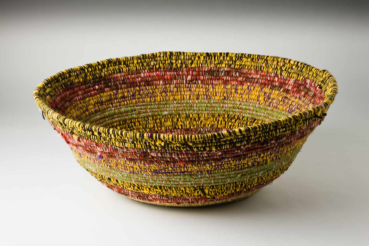 An oval coiled multicoloured yarn and plant fibre basket. The plant fibre in the centre of the basket is covered by purple-yellow yarn, followed by horizontal alternate yarn coverings over the fibre. The yarn colours are green-yellow, tan-yellow, purple-red, yellow-black, green-silver-brown and red-white-brown. The top plant fibre is covered by yellow-black yarn. - click to view larger image