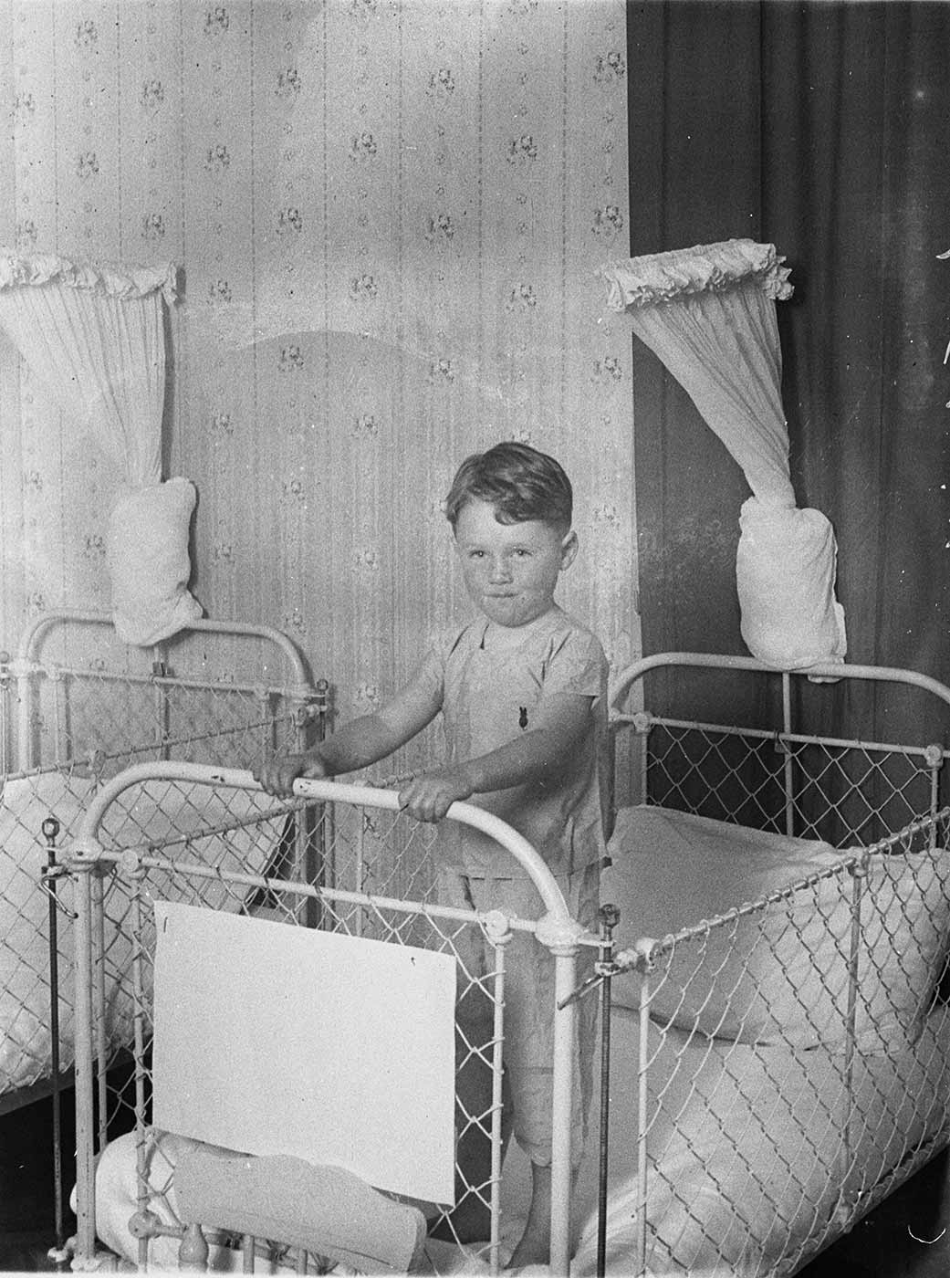 Boy standing in a metal framed cot with wire mesh sides. - click to view larger image
