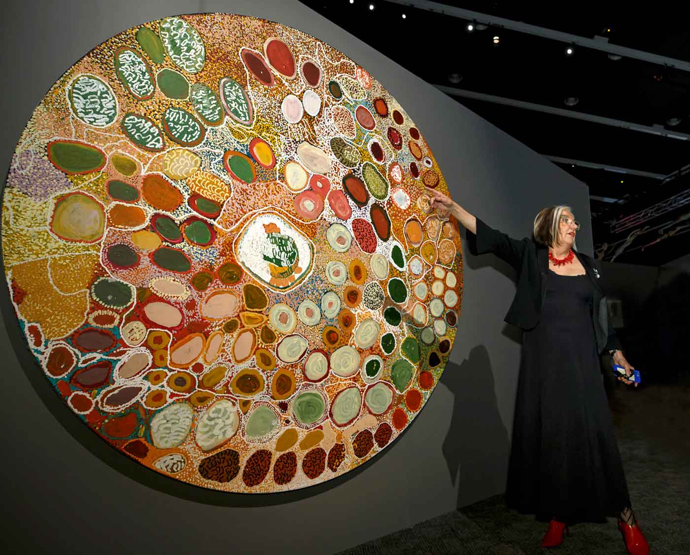 A woman pointing at a large circular Indigenous artwork mounted on an exhibition panel.