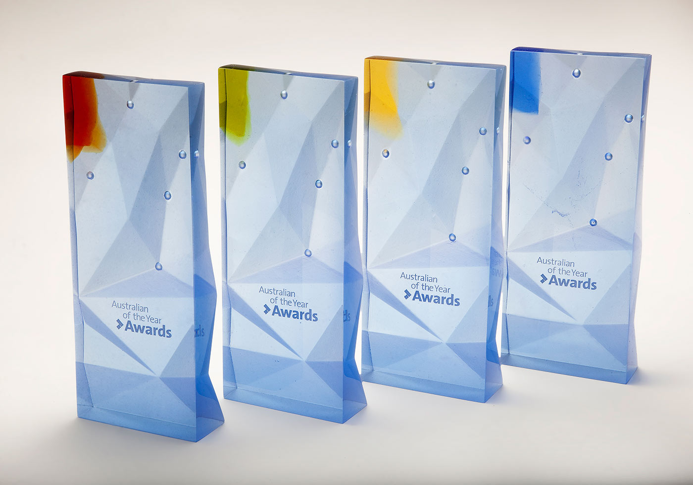 Four largely clear glass trophies with text 'Australian of the Year' and a small coloured glass tab top left. - click to view larger image