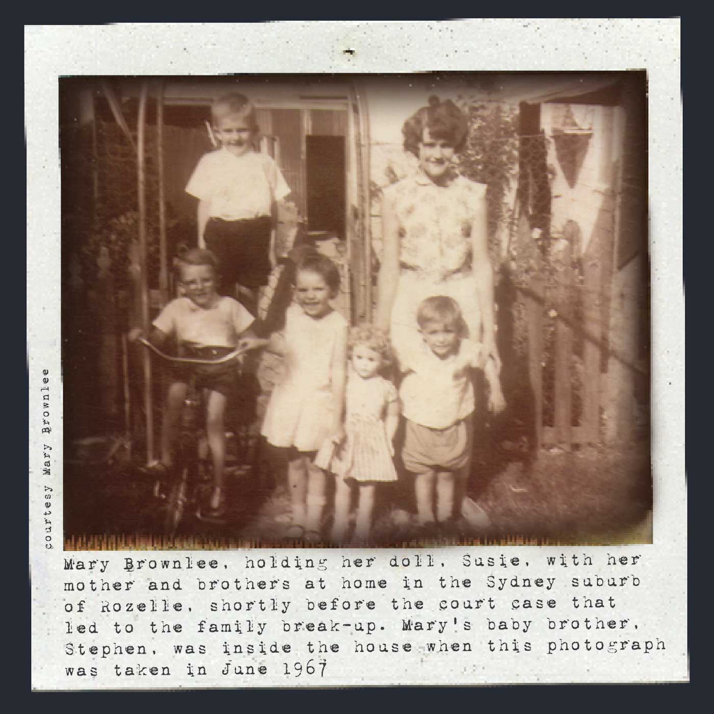 Black and white Polaroid photograph showing a young woman with three small boys and a small girl outside a house. They are positioned in a group facing the camera and all smiling. One the boys is sitting on a tricycle and the girl is holding a large doll. Typewritten text underneath reads: 'Mary Brownlee, holding her doll, Susie, with her mother and brothers at home in the Sydney suburb of Rozelle, shortly before the court case that led to their family break-up. Mary's baby brother, Stephen, was inside the house when this photograph was taken in June 1967'. In smaller text, on the left-hand side of the image, in a vertical direction, reads: 'Courtesy Mary Brownlee'. - click to view larger image