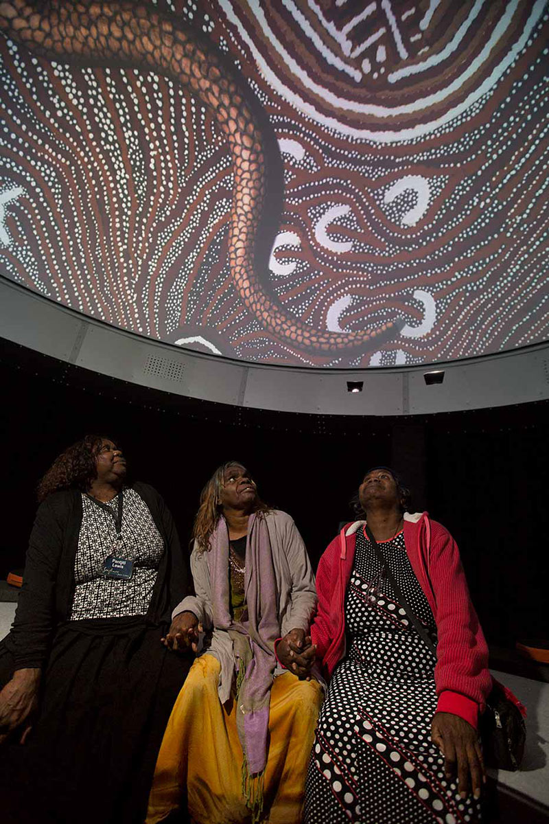 Three women sit hand in hand looking up at a digital screen featuring a serpent superimposed over artwork. - click to view larger image