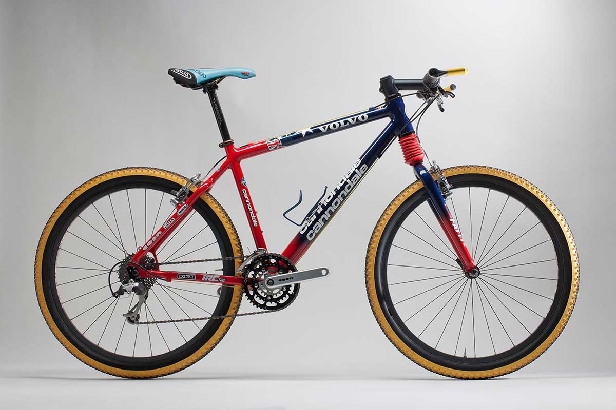 Front left view of a mountain bike with yellow-rimmed tyres. The bike has a pale blue seat and the frame is primarily painted red and dark blue, and the seat is pale blue. The bike frame has several stickers including 'Cannondale' and 'Volvo' on the main frame and 'Fatty' on the front forks.